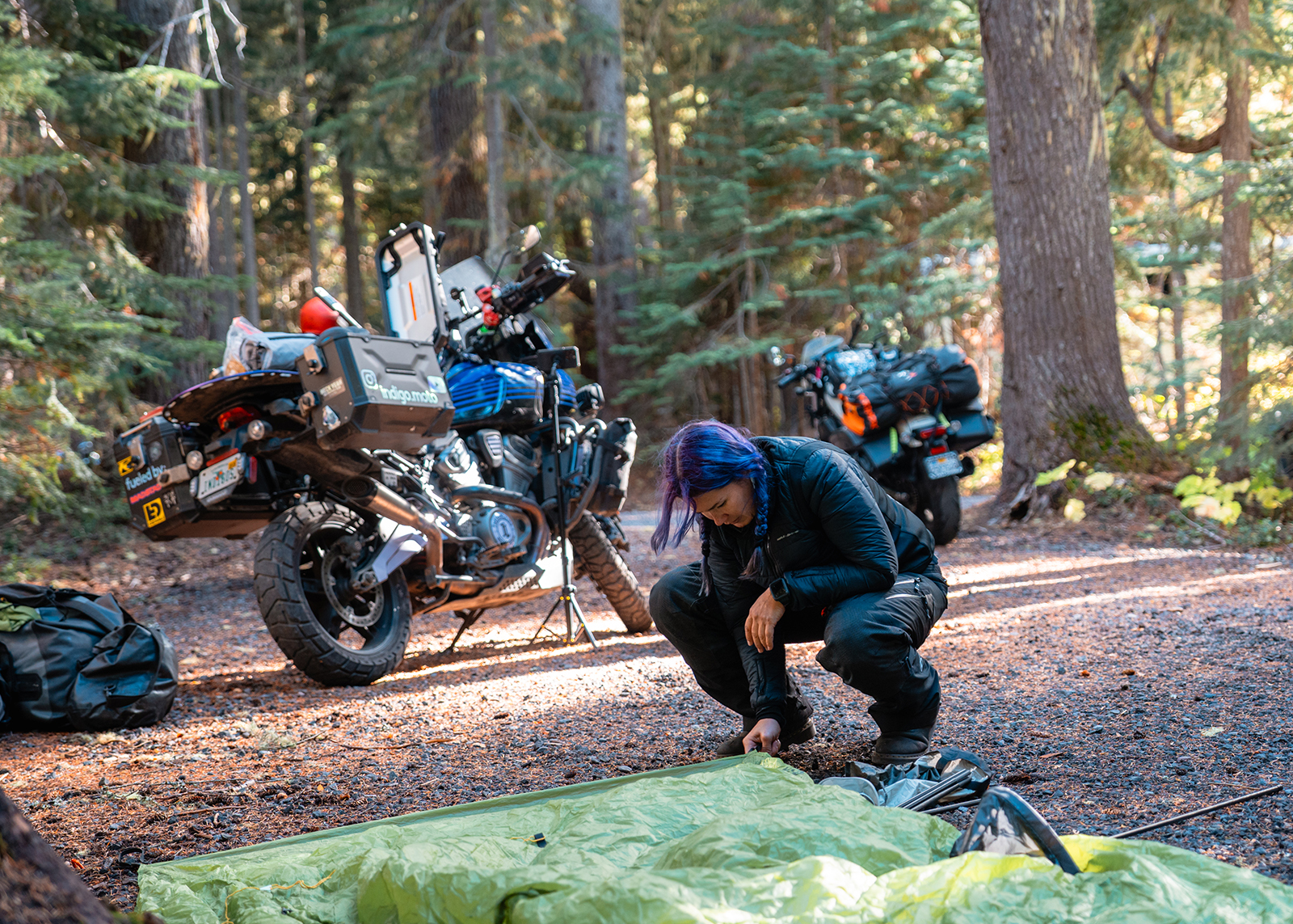 A rider sets up camp beside packed motorcycles in a shaded forest clearing.