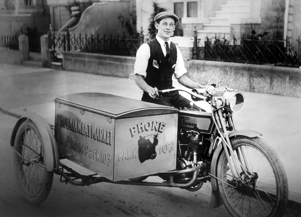 Vintage black-and-white image of an early delivery motorcycle driven by a man in a white shirt, vest, and tie