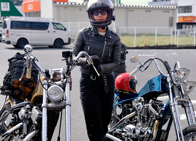 Adri Law standing between two parked choppers with helmets and bags visible on a paved street with buildings behind