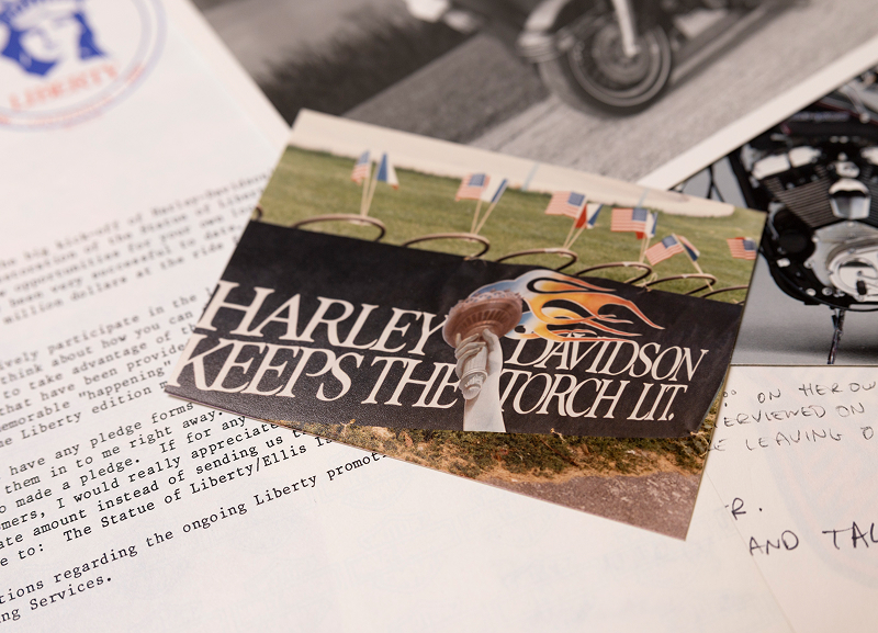 Printed card reading “Harley-Davidson Keeps the Torch Lit” resting on typed pages and motorcycle photos
