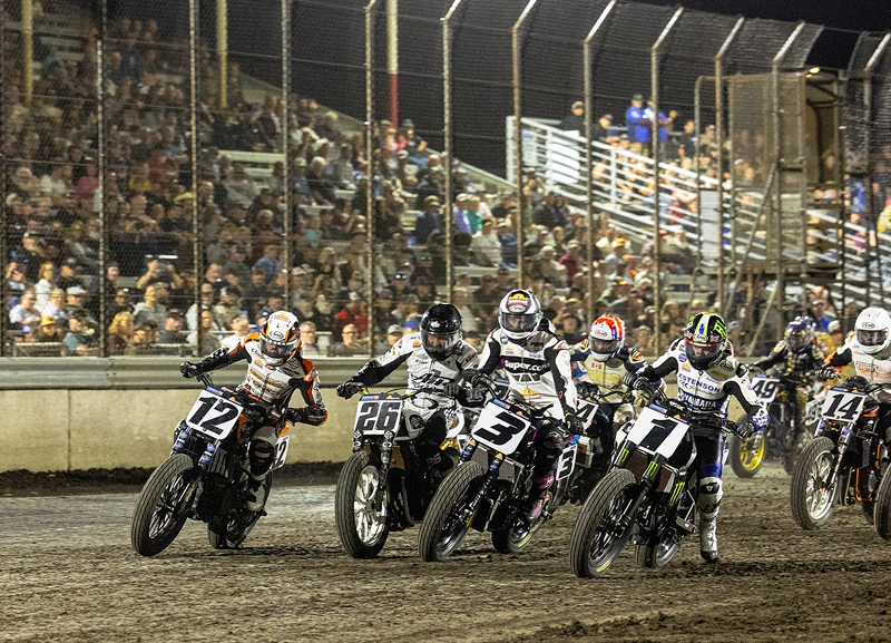 Flat track riders charge into first corner in tight pack kicking up dirt under stadium lights with full grandstands