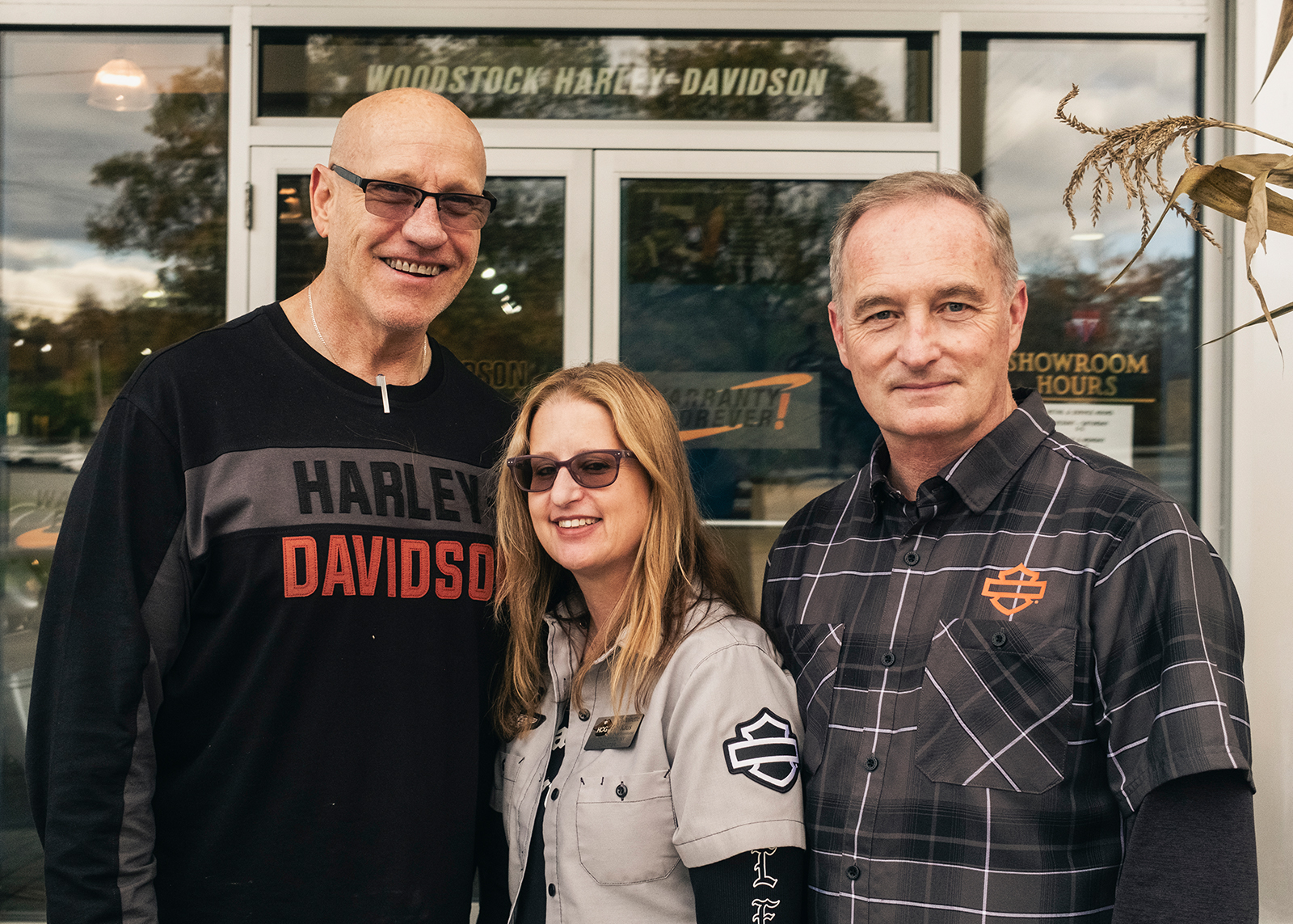 Three Woodstock Harley-Davidson employees pose in front of the entrance to the dealership.