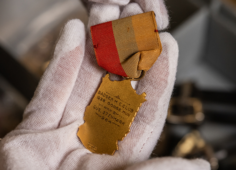 Back side of a gold medal with red ribbon, with engraved text saying “Badger MC Club 1936 Goose Run Won By Gus Stenmark”