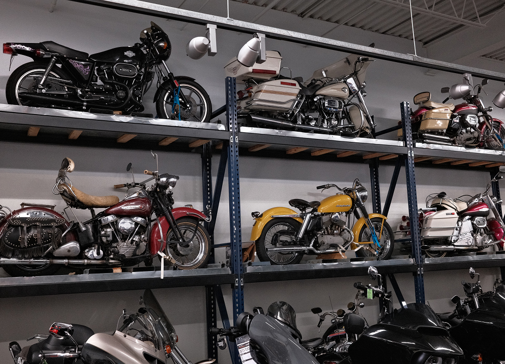 Rows of vintage H-D motorcycles stored on industrial shelving inside service area with various classic models