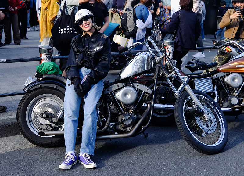 Shino seated on a custom motorcycle parked on a city street with a crowd gathered behind and additional bikes nearby