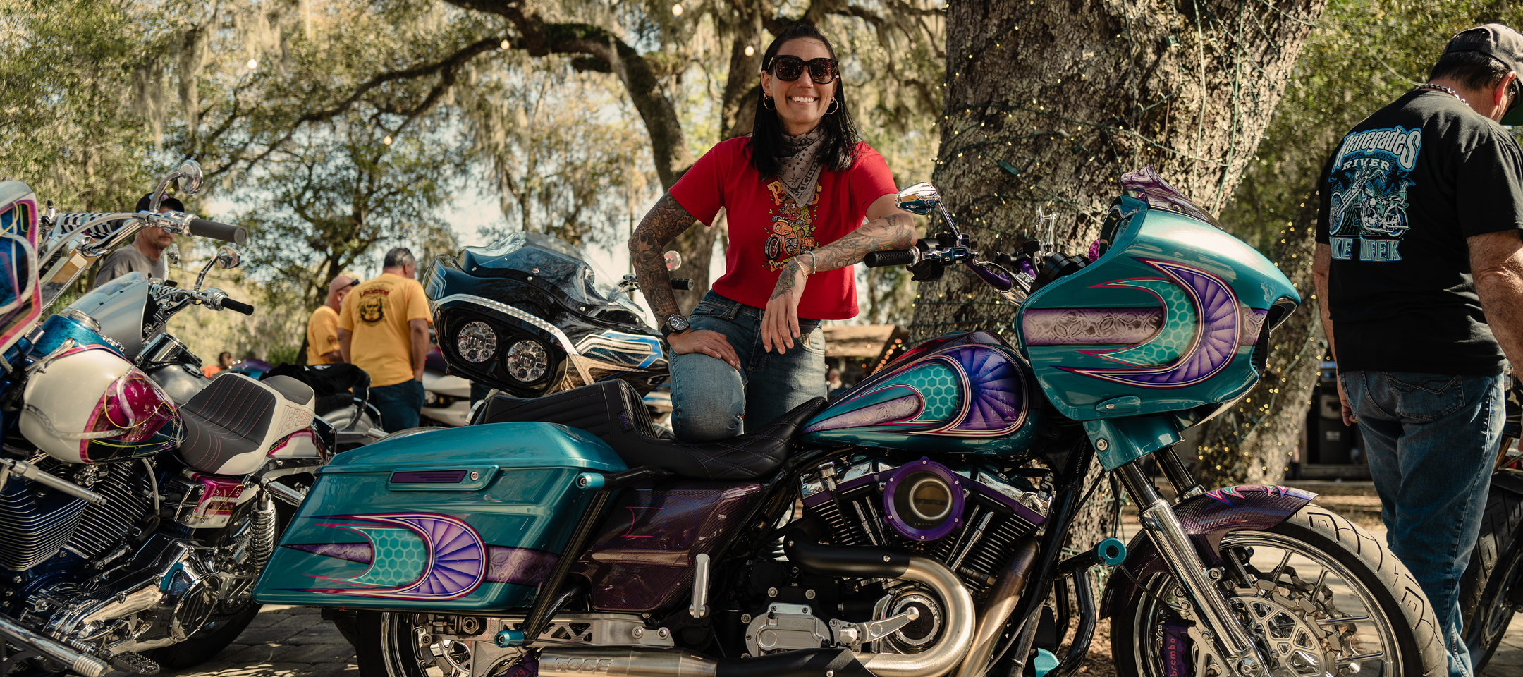 Jody Perewitz poses and smiles with a custom-painted touring motorcycle parked under trees