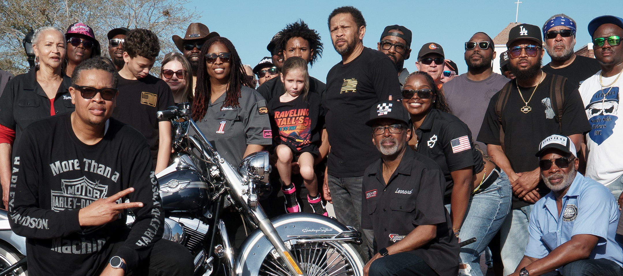 Group gathered outdoors around a classic Harley-Davidson motorcycle for a photo, wearing branded shirts and caps on a sunny day