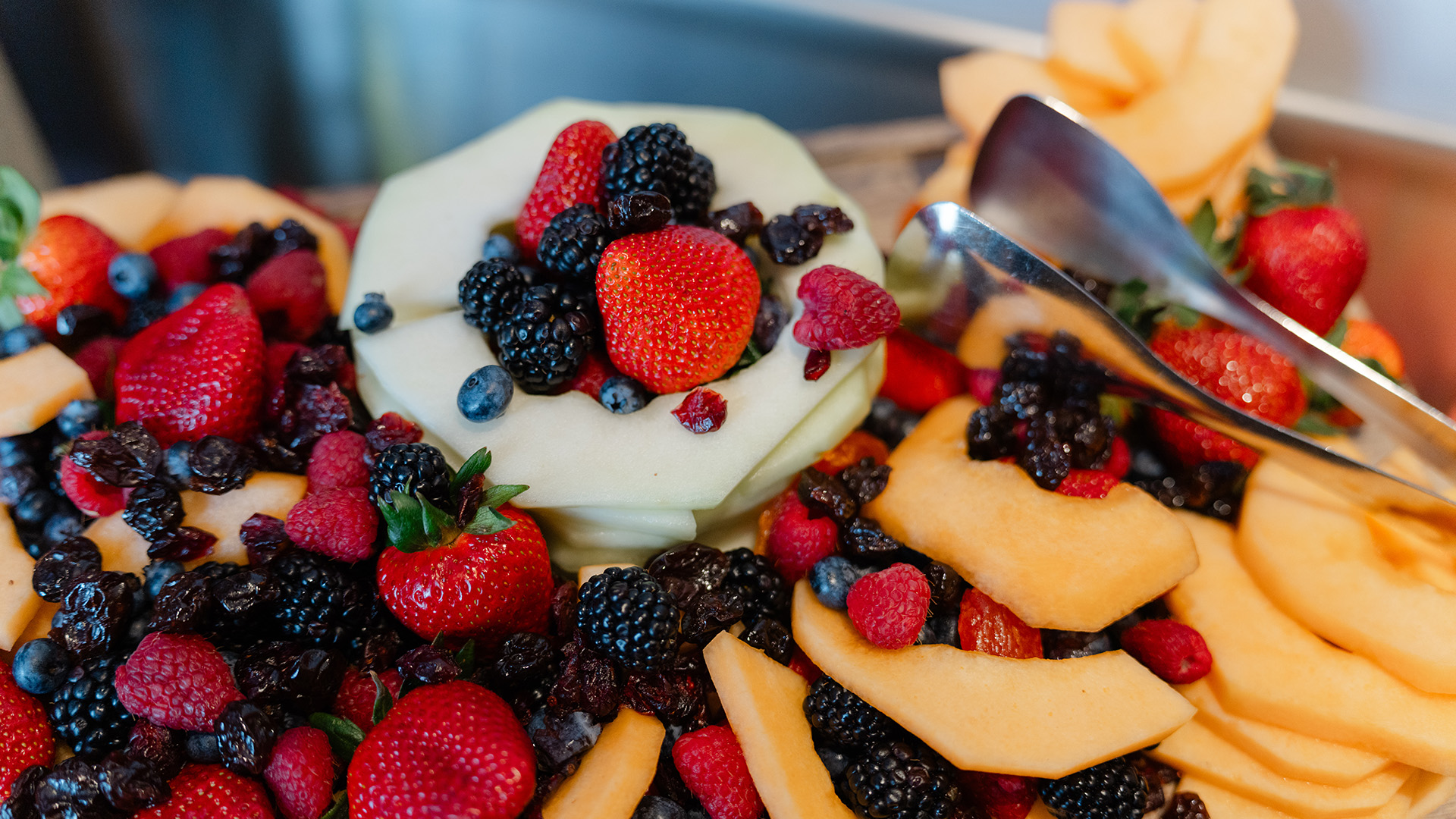 Fresh fruit display with berries, melon slices, and serving tongs