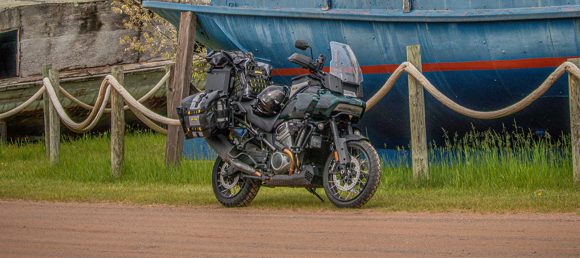 The author’s Harley-Davidson Pan America motorcycle loaded with gear parked on grass in front of a large weathered blue boat hull.