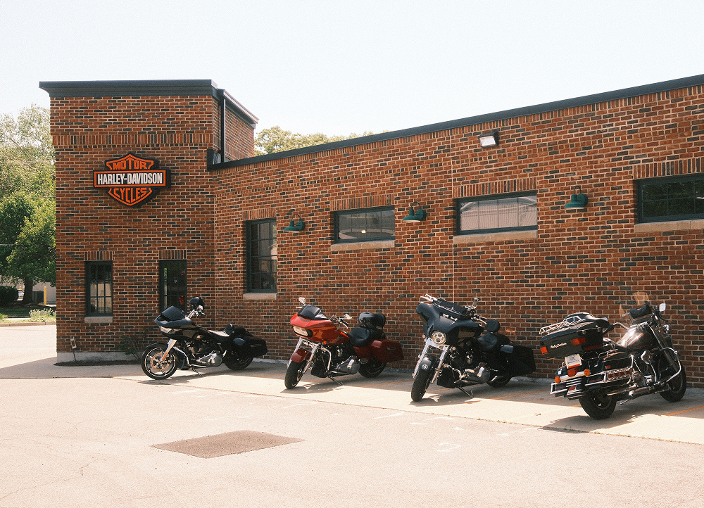 Row of parked Harley-Davidson motorcycles outside brick dealership with logo sign and patio seating