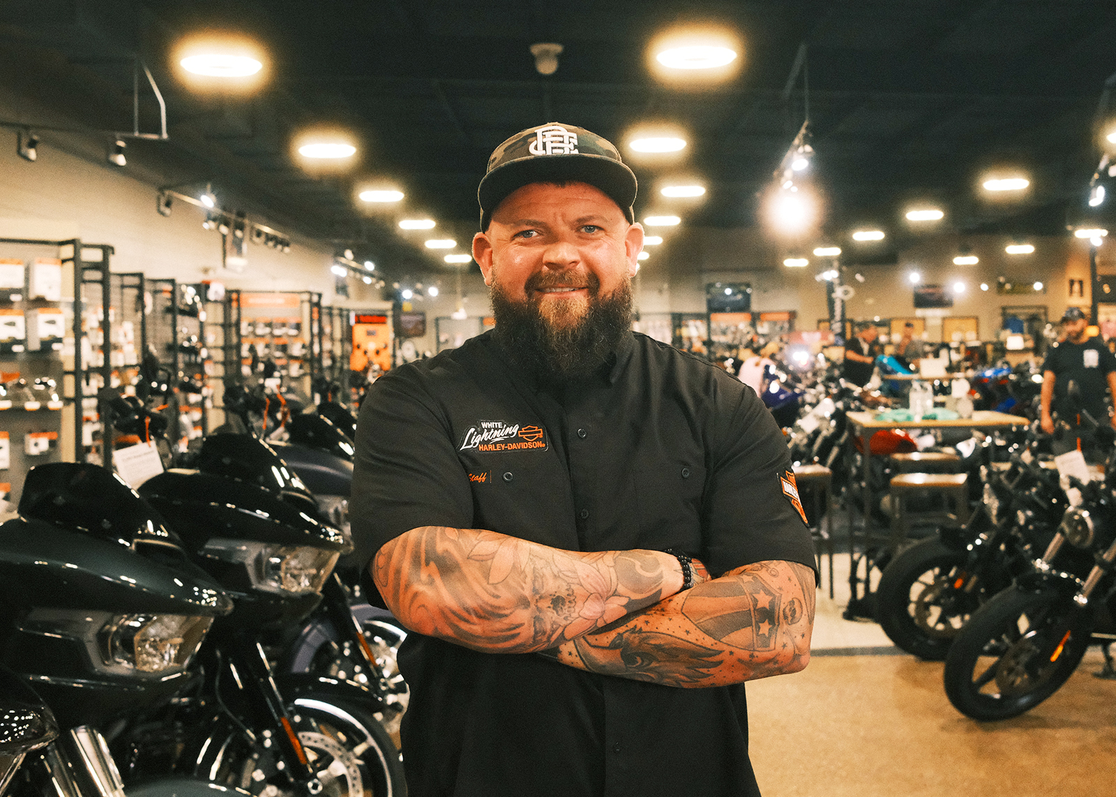 A portrait of Shawn Wells, the White Lightning Harley-Davidson General Manager, smiling with his tattooed arms crossed, standing amongst motorcycles on the showroom floor.