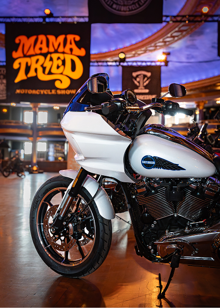 White custom motorcycle displayed under warm lighting with large event banners in the background