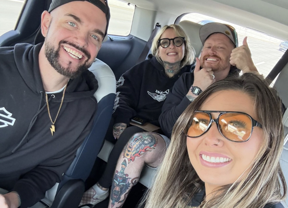 Four friends take a selfie inside a vehicle during a road trip, with smiles, sunglasses, and tattoos visible