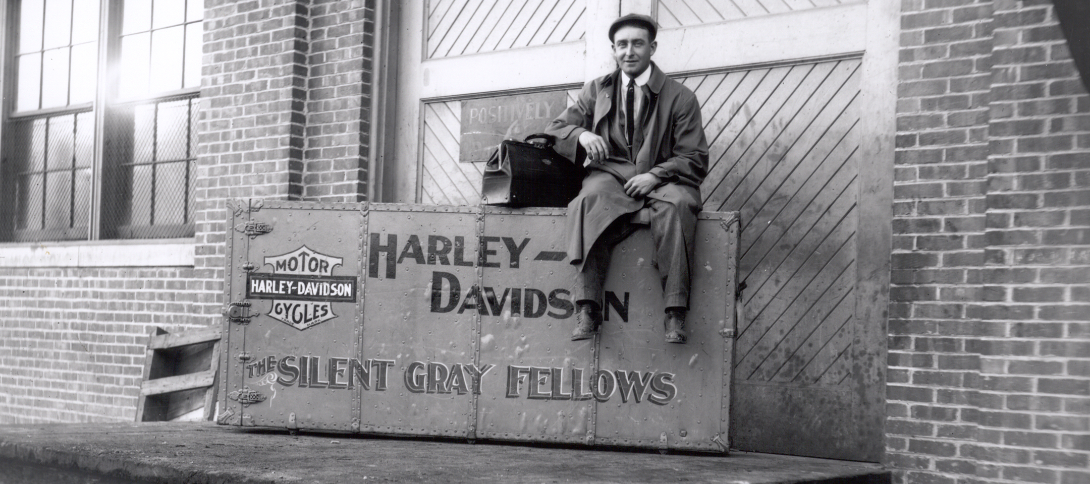 Vintage photo of a man sitting on an historic Harley-Davidson shipping crate with early Bar & Shield logo outside a factory warehouse building