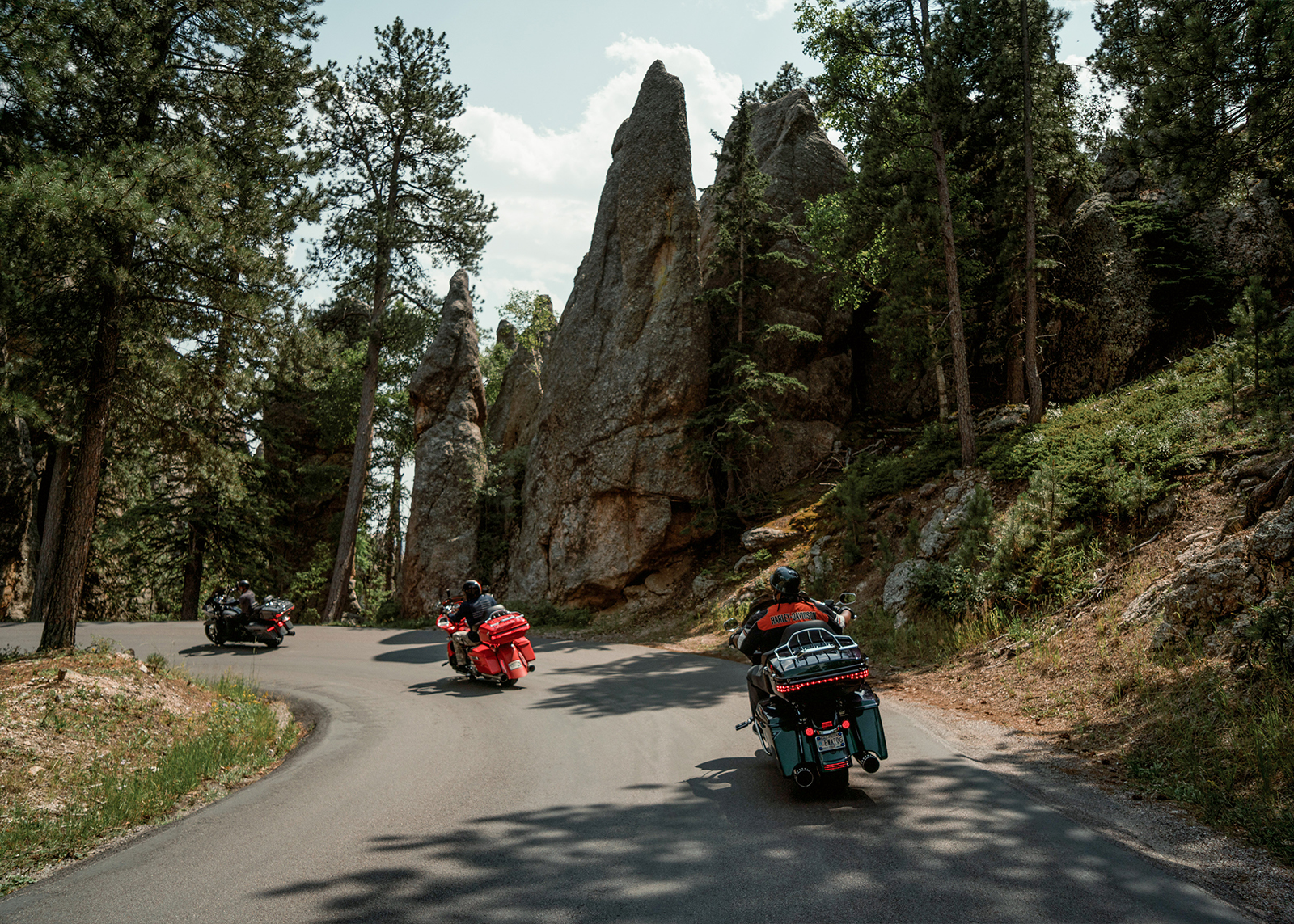 3 motorcycles go around a corner past tall, sharp rock formations on the Needles Highway in South Dakota.
