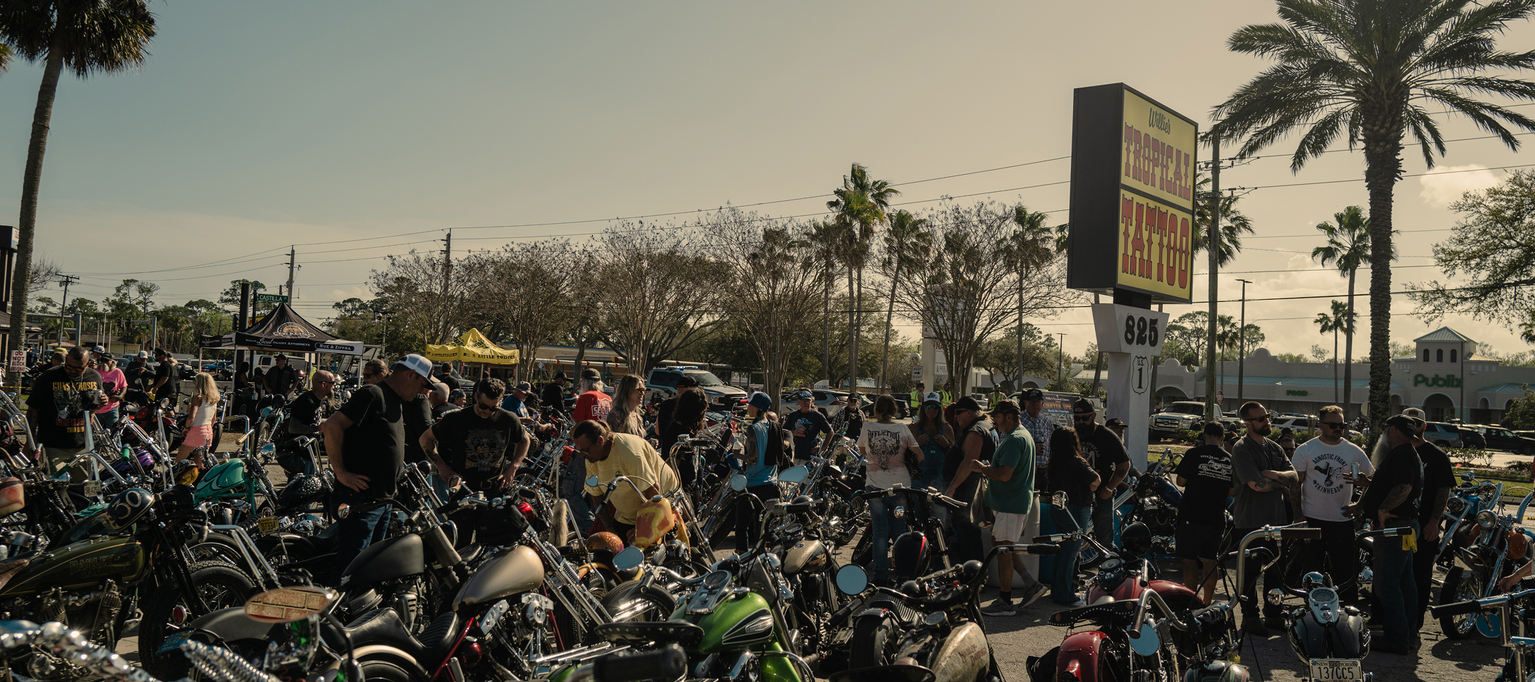 Crowded motorcycle gathering with rows of parked bikes and attendees outside a palm-lined roadside venue