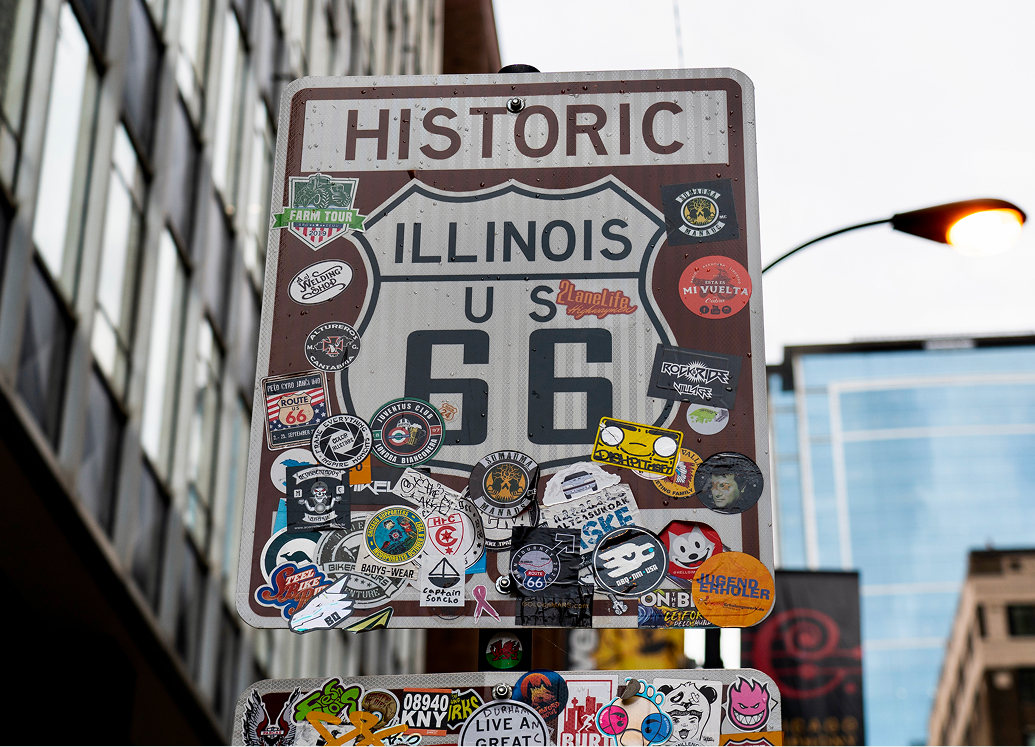 Historic Illinois Route 66 sign covered in colorful stickers, with city buildings rising in the background