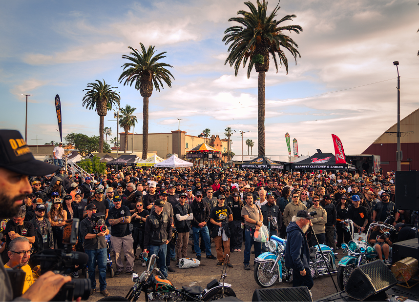 Large crowd filling a palm‑lined street at an outdoor motorcycle show during sunset