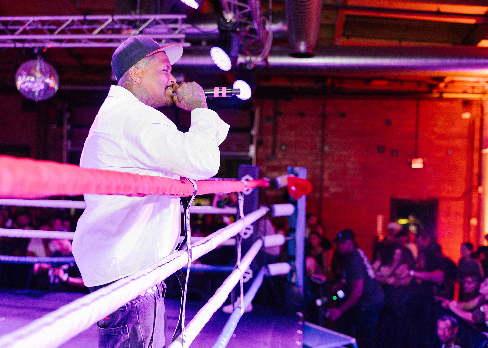 Rapper YG in a white long sleeve shirt and baseball hat performs to a crowd from the inside of a boxing ring.