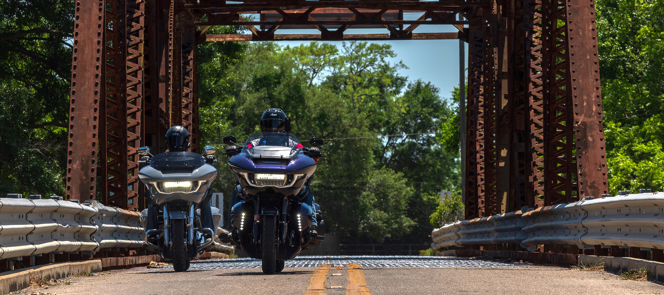  Two touring motorcycles ride side by side across a narrow steel bridge, framed by rusted trusses and trees
