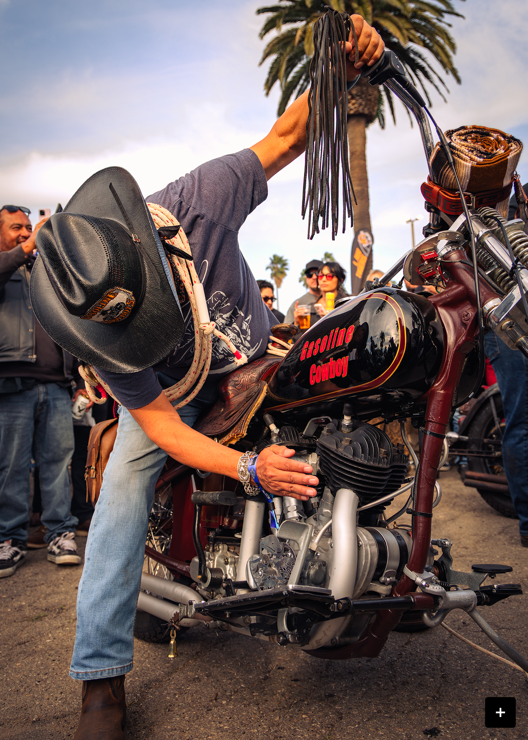 Person tuning a vintage motorcycle outdoors as others watch during a motorcycle gathering