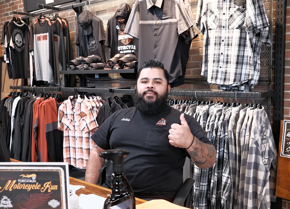 Dealership employee giving thumbs-up behind counter with apparel racks and Harley-Davidson clothing displays