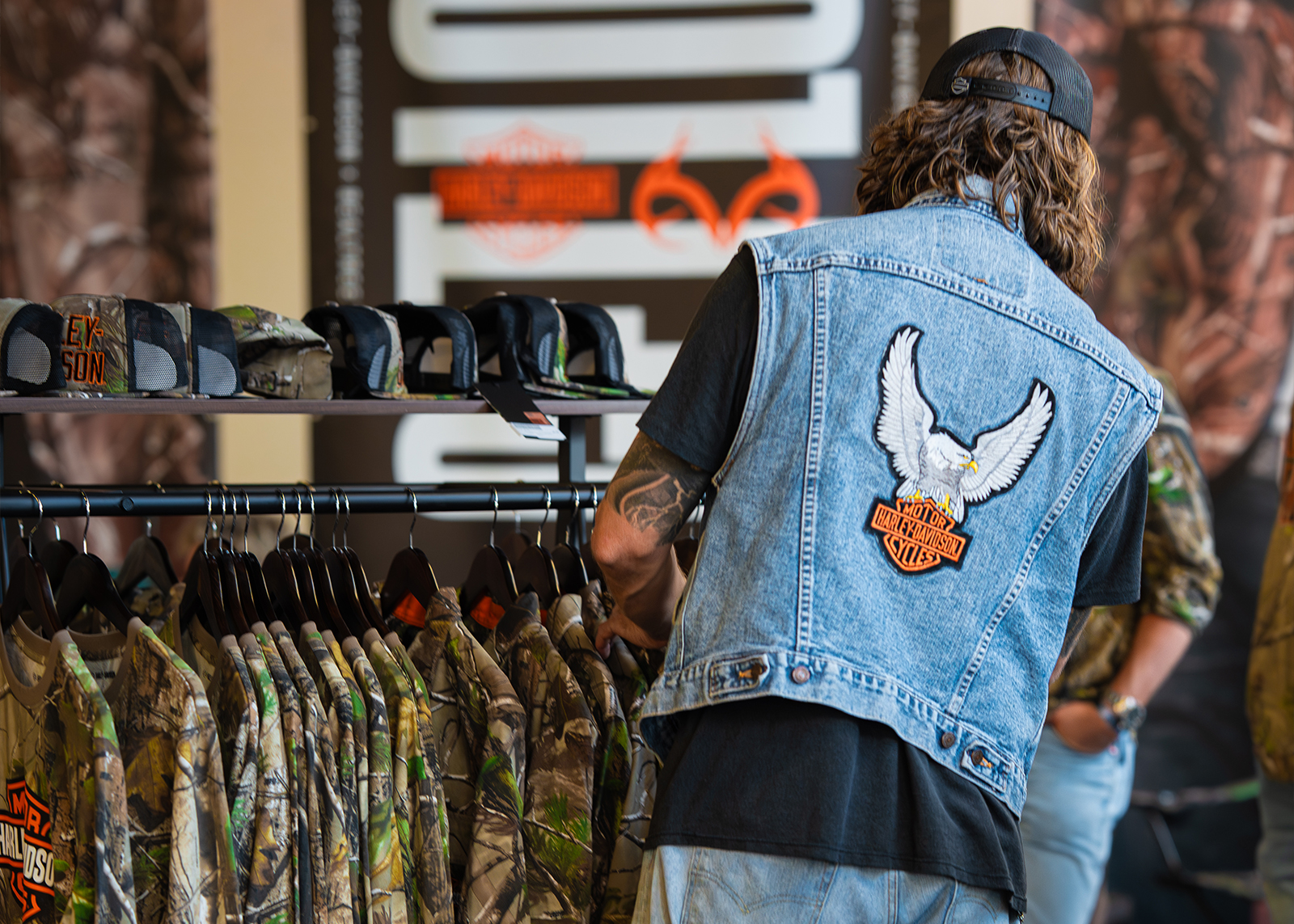 Rider in denim vest browses Harley-Davidson x Realtree camo apparel and hats on display at The Congregation Vintage Motorcycle Show.