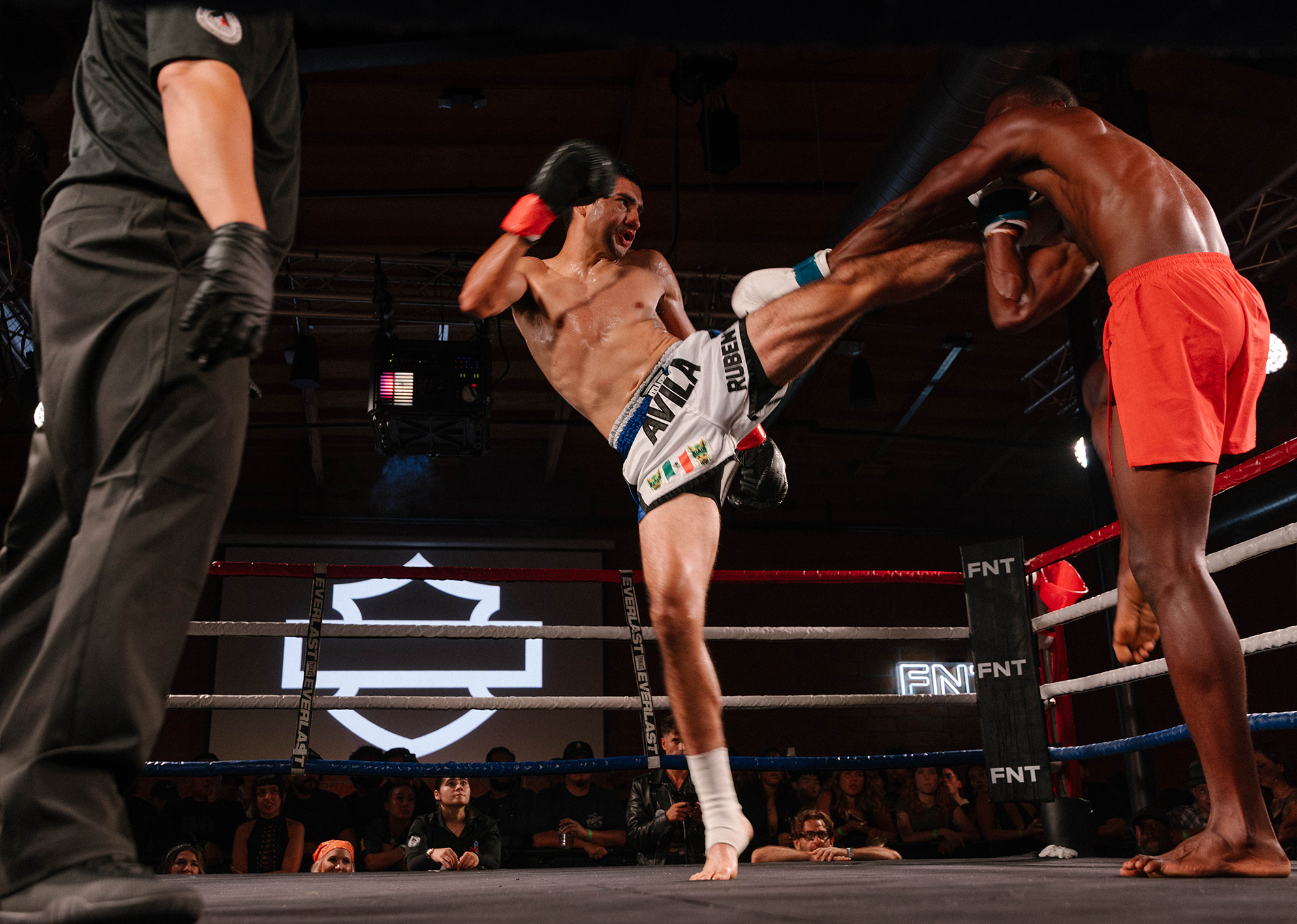 Fighter Ruben Avila in white shorts delivers a high kick to opponent Amor Valentine in red shorts during a match as referee looks on.
