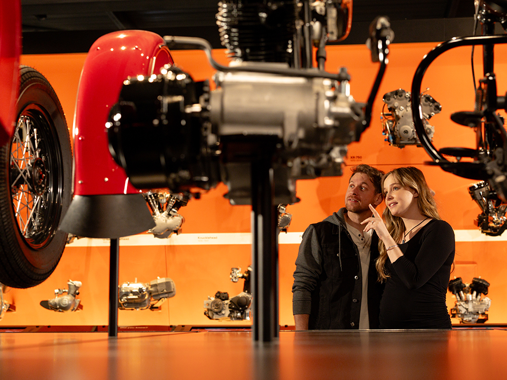 Visitors examining motorcycle engines and components displayed on wall