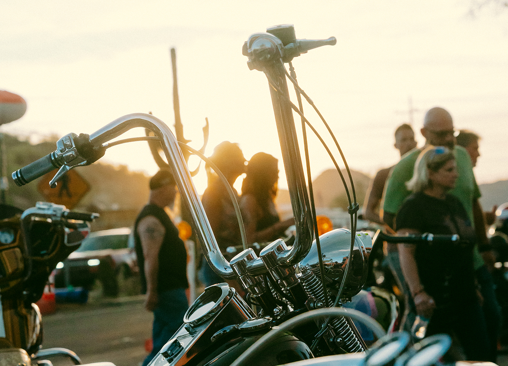 Tall chrome handlebars and front end of a custom motorcycle with people and setting sun in background