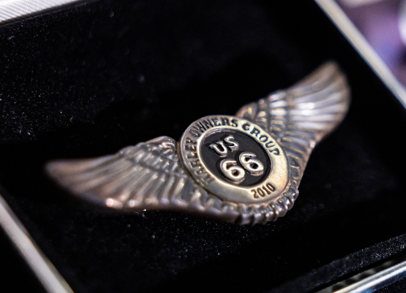 Close-up of a winged 2010 Route 66 badge pin with Harley Owners Group branding on a dark background