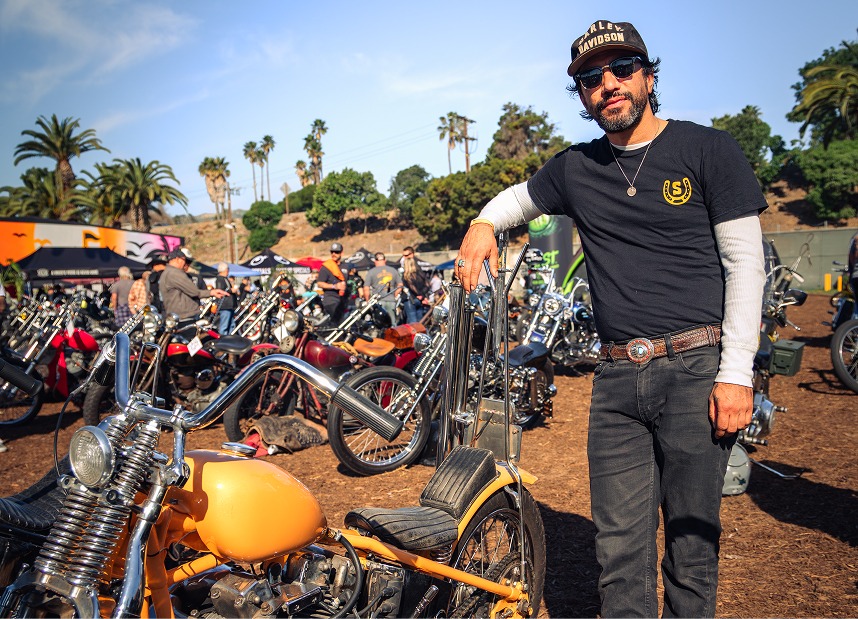 Person standing beside custom motorcycles at an outdoor show lined with palm trees