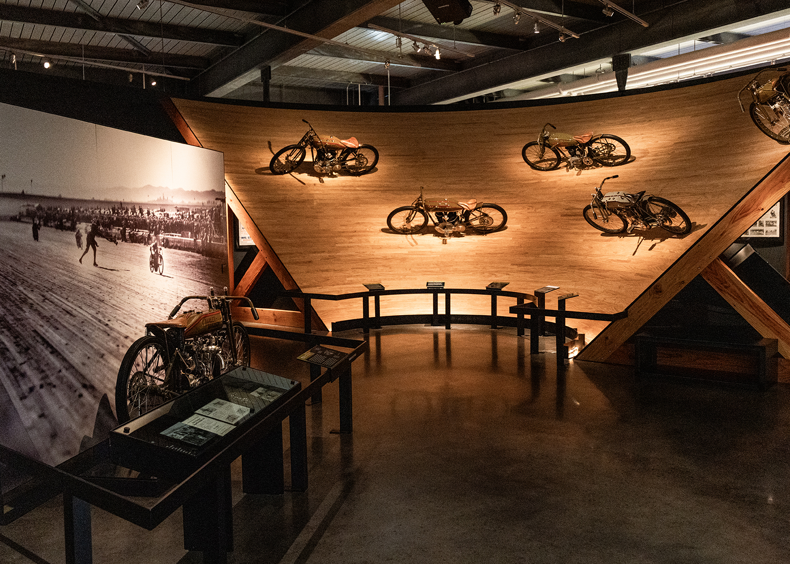Exhibit at the Harley-Davidson Museum featuring vintage motorcycles mounted on a curved section of board track with historic photos