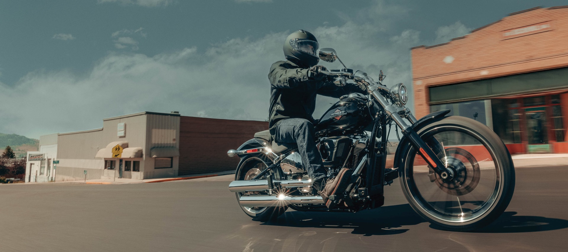 A motorcycle rider cruises through a small town on a 2026 H-D Breakout motorcycle as they pass by brick and storefront buildings.