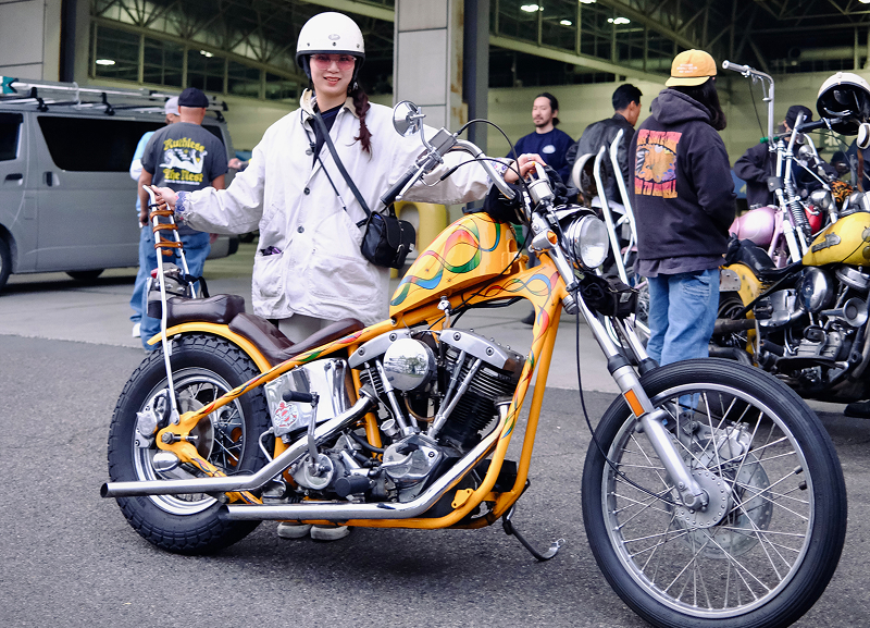 Riders gathered with parked custom motorcycles in an urban lot with one rider standing behind a bright orange bike