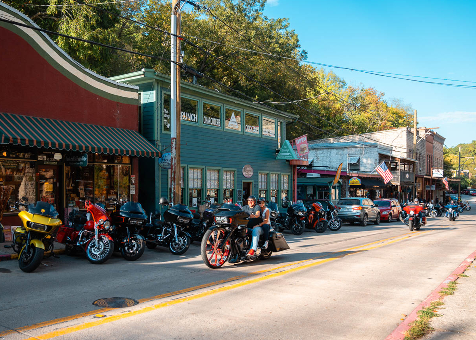A line of bikes is parked in front of small businesses while other Harley-Davidsons ride by 