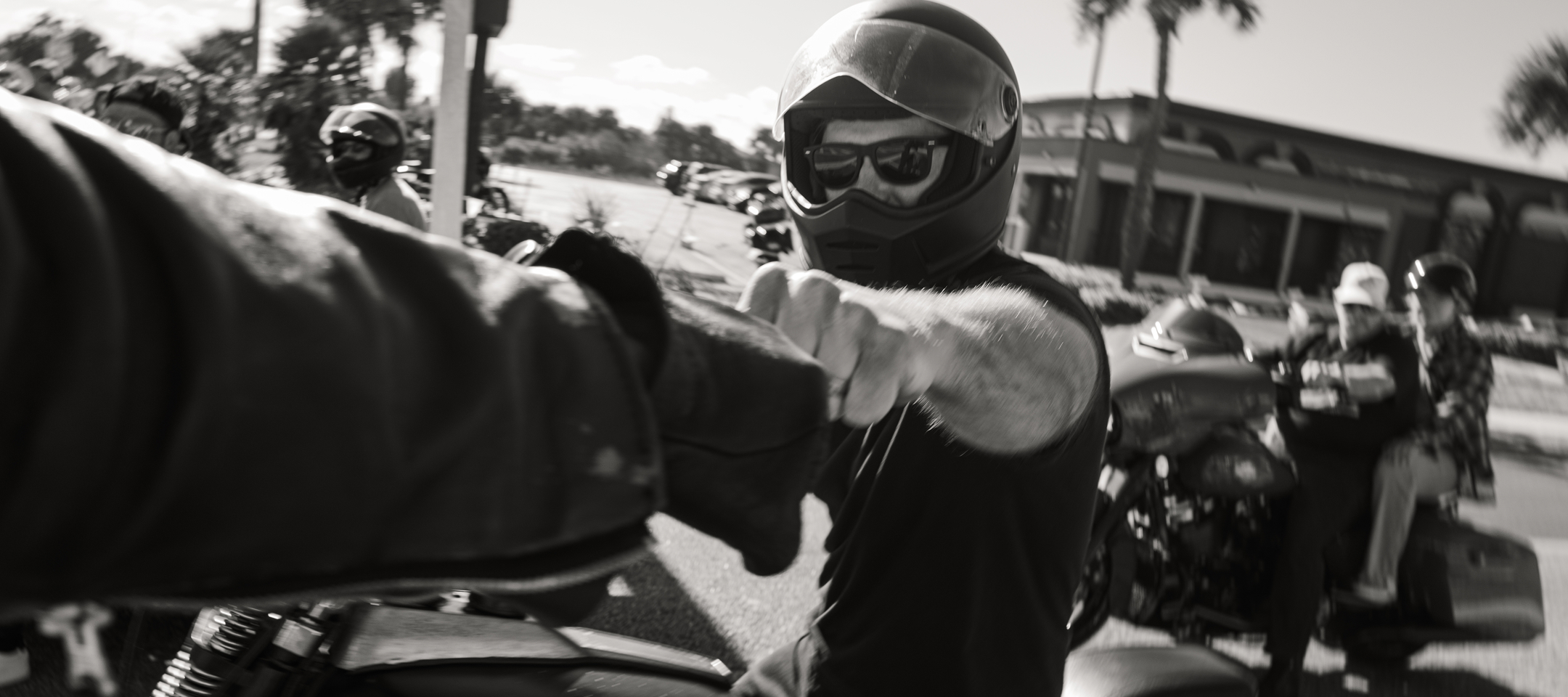 Helmeted motorcyclist reaching out to bump fists with another rider while a group rides together on a city street