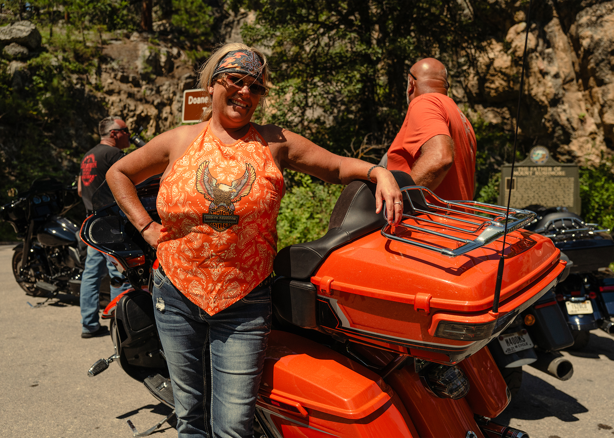 Smiling woman in orange Harley-Davidson top leans on matching orange touring motorcycle.
