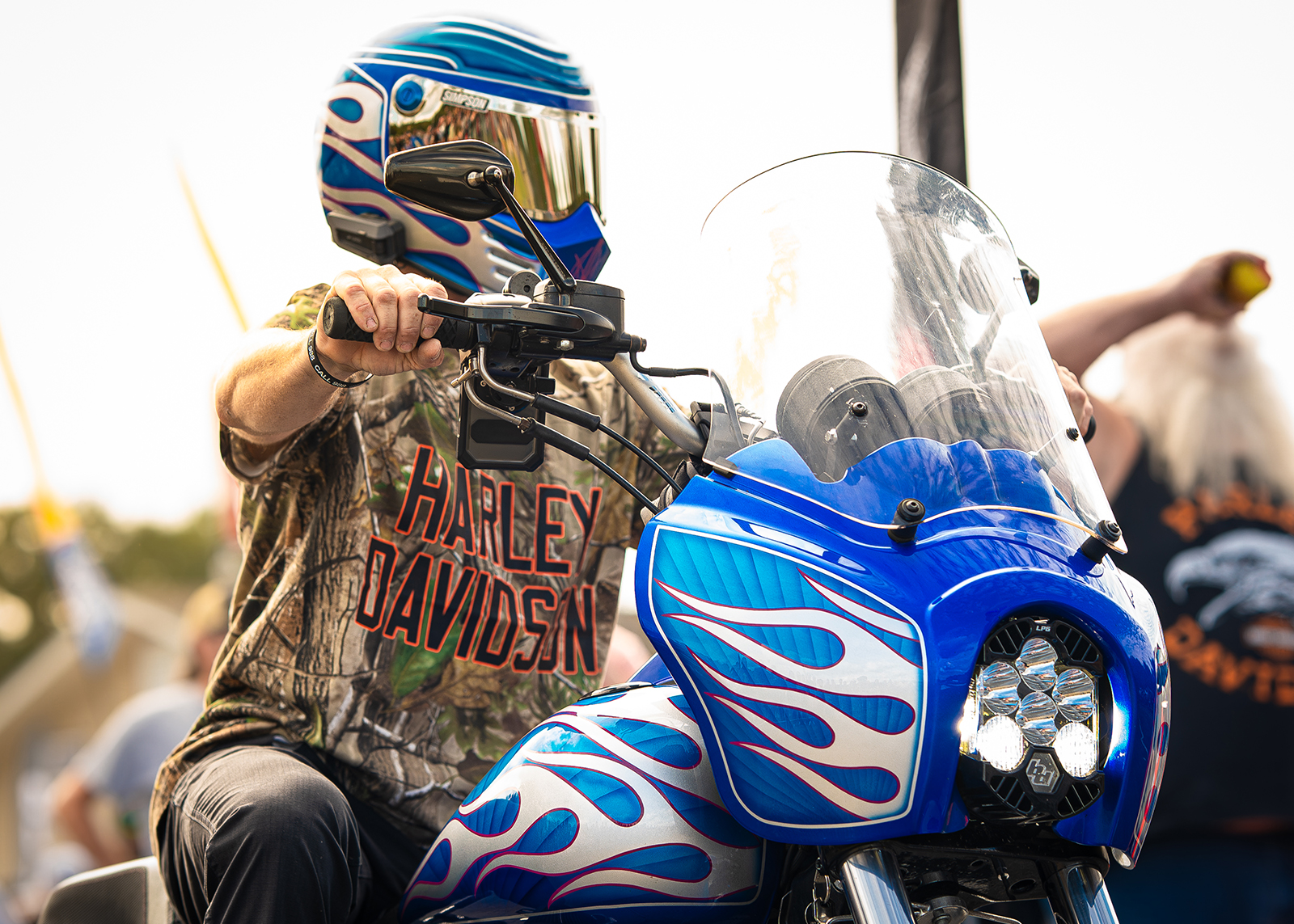 Rider in Harley-Davidson X Realtree shirt and blue flame helmet grips the handlebars of a custom motorcycle.