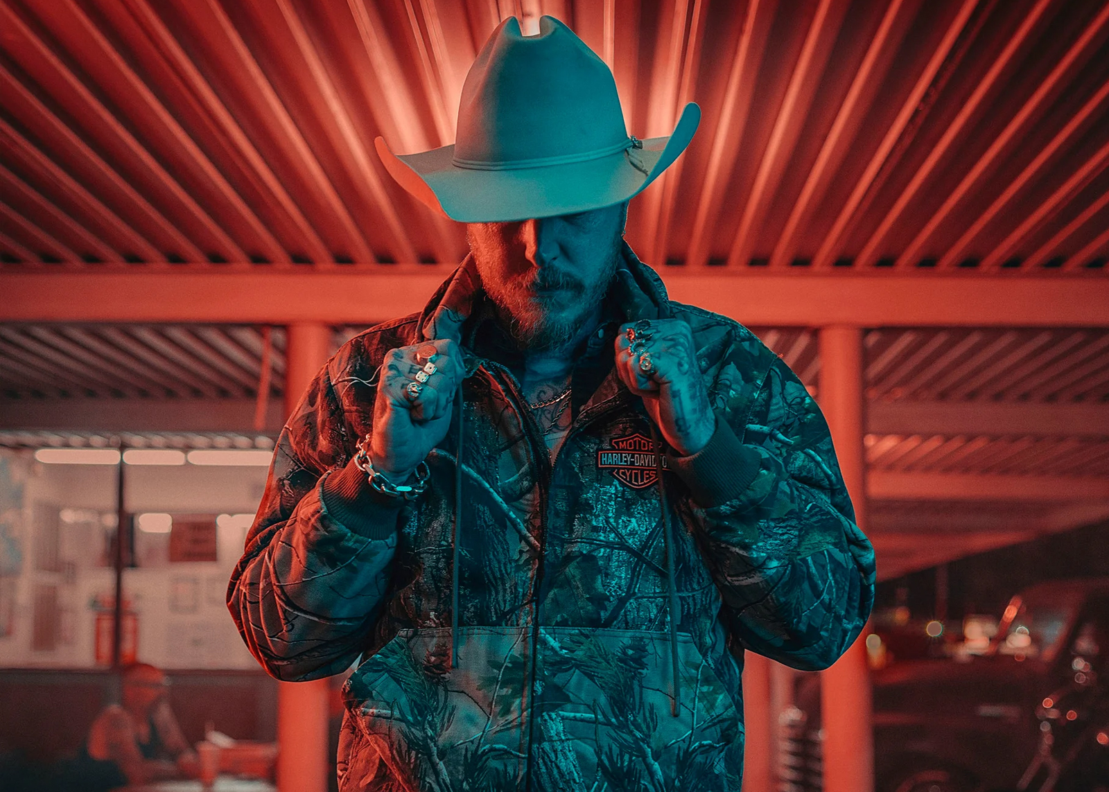 Man in cowboy hat and camo Harley-Davidson x Realtree hoodie stands in dramatic red and blue lighting at night.