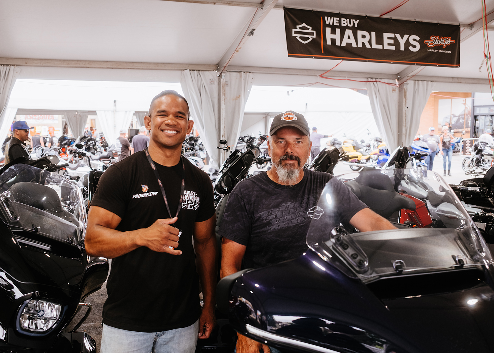 A smiling staff member and customer standing by a motorcycle under a large tent at Sturgis Harley-Davidson.