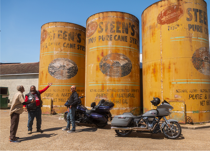 Motorcycles parked in front of large yellow industrial tanks with riders standing nearby outdoors