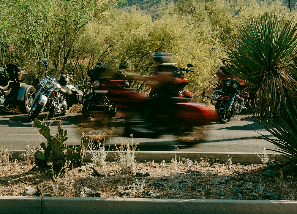 Motorcycle riding past desert landscaping and parked bikes, long exposure creating motion blur of the rider.
