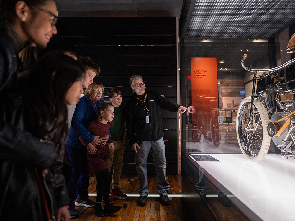 Guide explaining vintage motorcycle display to group of visitors in museum