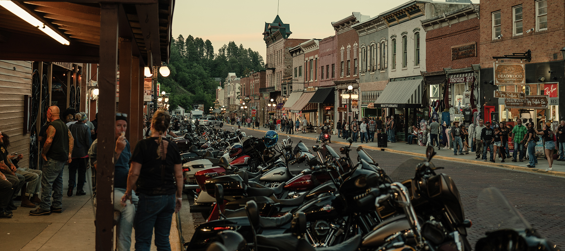 Motorcycles line a bustling Deadwood street as people stroll past historic brick buildings.