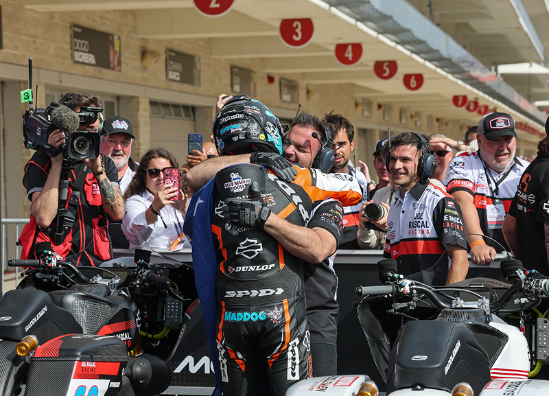 Rider celebrates in pit lane while surrounded by crew members, photographers, and team equipment