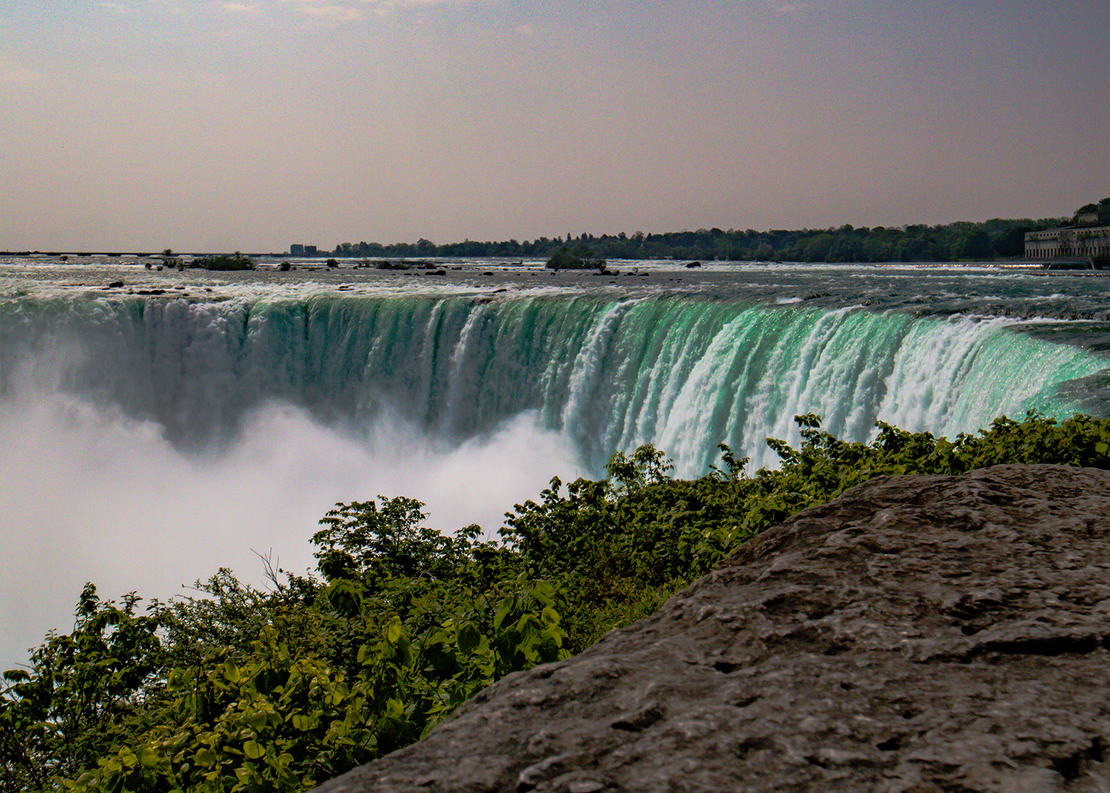 Massive turquoise water cascades over Niagara Falls, mist rising as greenery frames the powerful scene.
