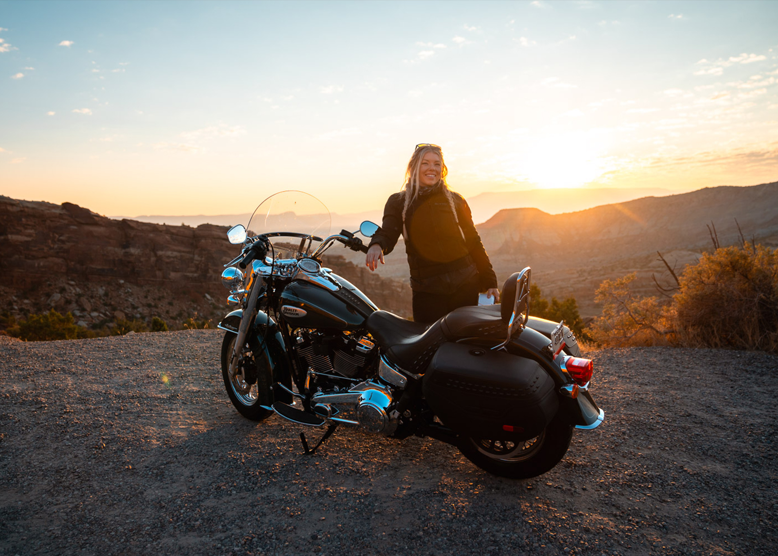 A woman smiles standing behind her parked motorcycle at sunset