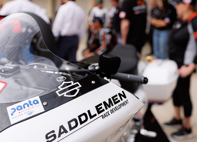 Close-up of a race motorcycle fairing with Saddlemen branding with crew and equipment blurred in the background