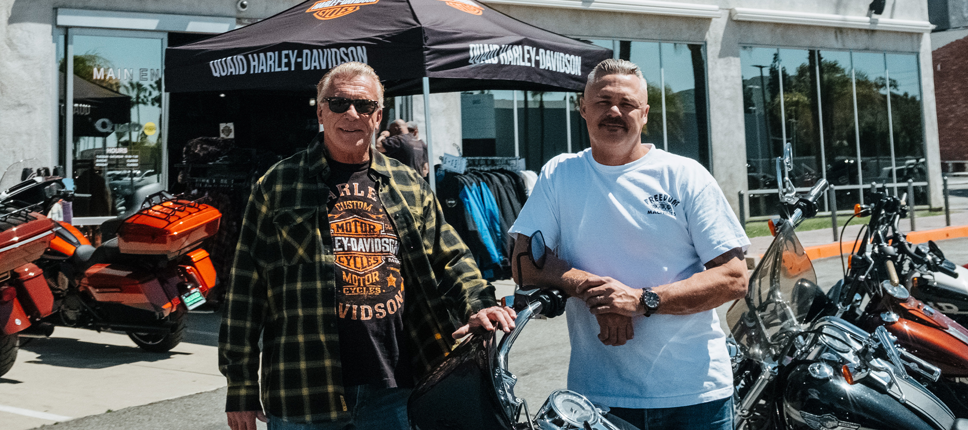 Two members of Quaid staff pose with motorcycles in the parking lot in front of the Quaid Harley-Davidson dealership.